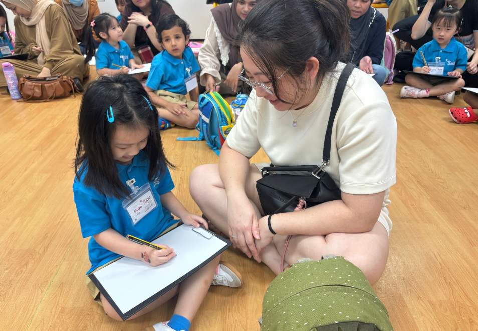 Girl in blue uniform writes on a clipboard while a seated woman watches; other children sit behind.
